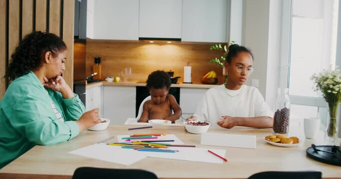 A mother with two children has breakfast cereal sitting at the table at home. A naked baby is sitting in a high chair