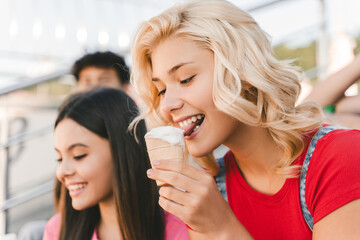 Cute smiling teenage girl eating tasty ice cream sitting on the street with friends, having fun. Friendship, positive lifestyle, summer concept