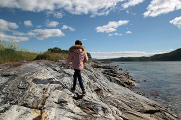 A girl in a pink jacket walks along a rock near the sea, a teenager walks on the shore