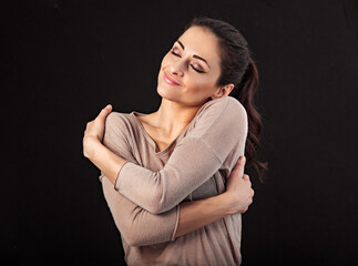Beautiful happy calm smiling woman hugging herself with love on studio black background. Closeup