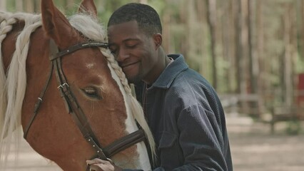 Medium shot of young black man petting beautiful horse with braided mane, smiling and bonding with her outdoors