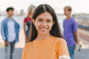 Portrait of smiling teenage girl wearing stylish yellow t shirt looking at camera standing on the street with friends on background. Summer, positive lifestyle concept 