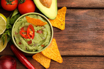 Bowl of delicious guacamole, nachos chips and ingredients on wooden table, flat lay. Space for text