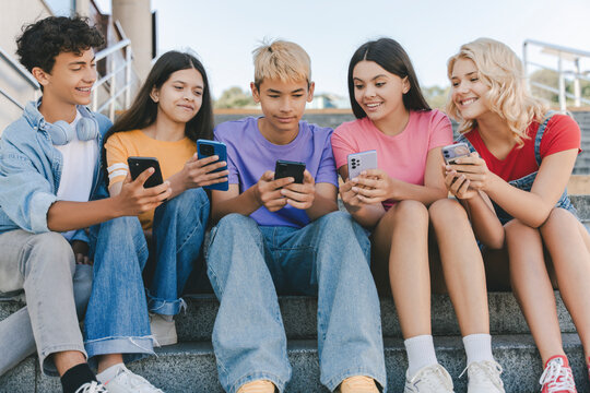 Group of smiling friends, multiracial teenagers holding mobile phones watching video, communication online, playing mobile game, talking sitting on stairs. Technology, social media concept