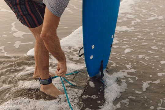 Male sportsman attaching leash of blue surfboard to ankle, preparing for activity standing on sandy beach by sea. - Powered by Adobe