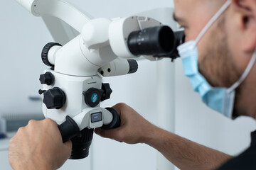 Male doctor in blue medical mask holding hands on handles of dental microscope and looking through binocular equipment.
