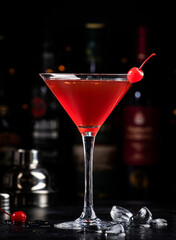 Manhattan alcoholic cocktail drink with bourbon, red vermouth, bitter, ice and cocktail cherry in glass, dark bar counter background, bar tools and bottles