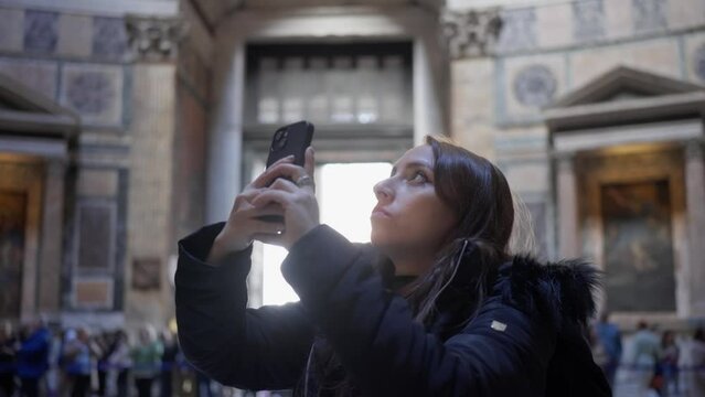 Side view of young Hispanic woman in warm jacket using phone to take photo of interior of Pantheon temple in Rome, Italy