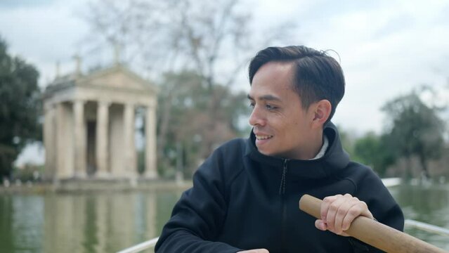 Smiling young Latin man in casual clothes rowing boat with oars in pond with lake near Temple of Aesculapius in gardens of Villa Borghese, Roma, Italy
