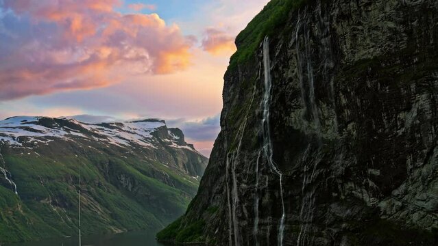 Fantastic picturesque silhouette view of steep snow-capped mountain cliffs with gorgeous waterfalls and dramatic colorful sunset sky on the Geiranger Fjord, Norway