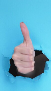 Vertical video. Like hand. Perfect choice. Accepting yes thumb up gesture through hole in breakthrough paper ripped wall on blue background with free space.