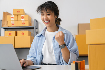 Startup small business, Young Asian woman using laptop with fists clenched celebrating victory expressing success and boxes for products to send to customers. working at the home office.