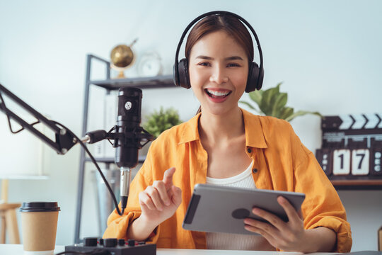Young Asian woman holding digital tablet and use microphones wear headphones with laptop record podcast interview for radio. Content creator concept.
