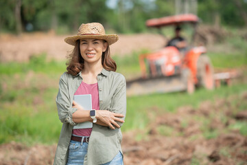 Attractive farmer, Young woman with laptop standing on field in sunset while tractor baling in...