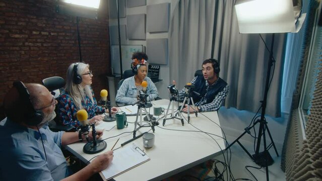 Group of senior and young podcast co-hosts sitting around the table in professionally equipped recording studio, talking in microphones, clapping hands and celebrating the last episode