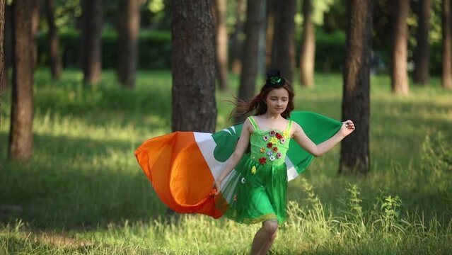 St Patricks Day celebration. Cheerful white girl in green pixie dress running in the wood with flag of Ireland in slow motion
