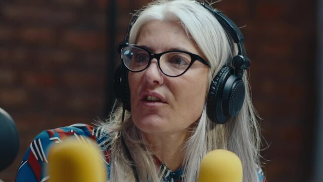 Beautiful senior woman with long gray hair wearing headphones speaking in microphone, interviewing guest, hosting podcast in the recording studio. Medium close-up shot