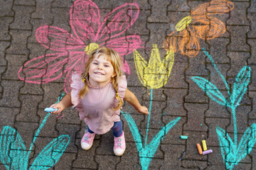 Little preschool girl painting with colorful chalks flowers on ground on backyard. Positive happy...
