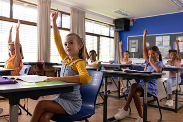 Diverse elementary schoolchildren raising hands at desks in class