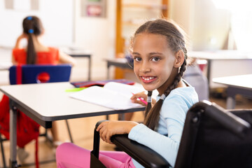 Portrait of happy diverse and disabled schoolgirls in wheelchair writing in classroom
