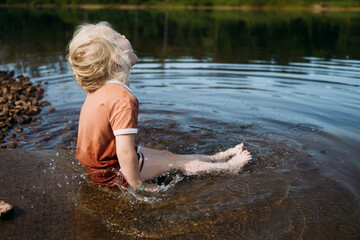 Child sitting in water in casual clothes at edge of pond splashi