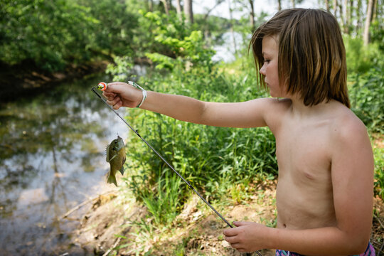 Young boy holding up a fish on fishing rod by a stream outdoors