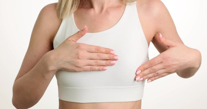 Female person in white top carefully touches chest to avoid pain standing on light background. Woman suffers from ache in breast. Breast cancer in women