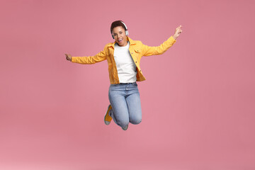 Happy young woman in headphones jumping while dancing on pink background