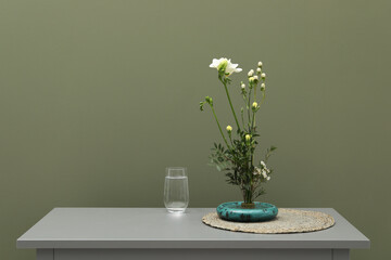Stylish ikebana with beautiful flowers, green branches and glass of water on gray nightstand near olive wall, space for text