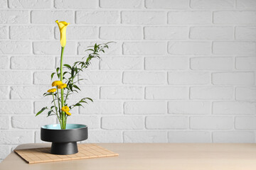 Beautiful ikebana for stylish house decor. Floral composition with fresh calla, chrysanthemum flowers and branches on wooden table near white brick wall, space for text