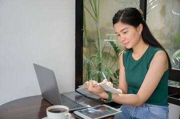 woman working freelance Young Asian woman sitting at work writing articles at wooden table at cafe with laptop and tablet watching webinar writing notes.