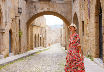 Fototapeta na wymiar summer trip to Rhodes island, Greece. Young Asian woman in ethnic red dress walks Street of Knights of Fortifications castle. female traveler visiting southern Europe. Unesco world heritage site.