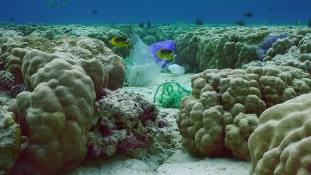 Closeup, Plastic pollution on beautiful shallow coral reef, Slow motion. Underwater landscape of shallow water coral reef polluted with plastic, tropical fish swims nearby. Camera moving forwards
