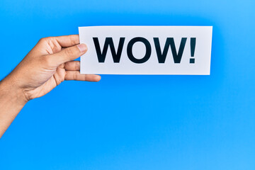 Hand of caucasian man holding paper with wow! message over isolated blue background