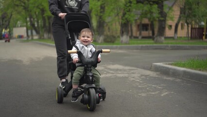 Obraz premium A young mother carries a small child on a tricycle. Two-year-old boy on a bicycle close-up.