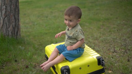 A little boy sits on a yellow suitcase in nature. A one-year-old child sits on a suitcase and rolls down.