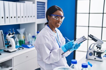 Young african american woman scientist smiling confident writing on document at laboratory