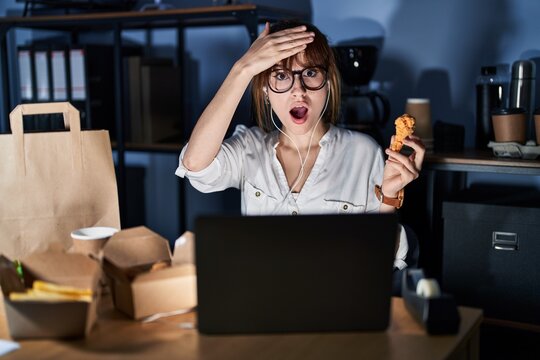 Young beautiful woman working using computer laptop and eating delivery food surprised with hand on head for mistake, remember error. forgot, bad memory concept.