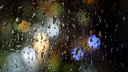 Closeup of rain droplets and streams flowing down the window at night. Colorful bokeh lights on the...