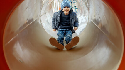 Cute smiling boy riding down in the tunnel slide at playground. Happy kids, fun outdoors, leisure in park.