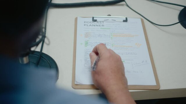 Male host sitting at desk in the recording studio, reading handwritten podcast planner and outlining task with pen. Close-up shot, over the shoulder view