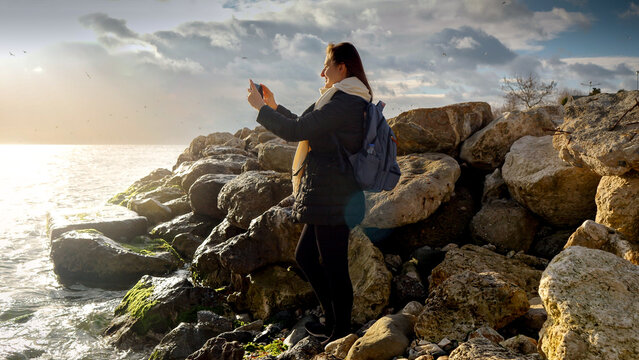 Beautiful young woman standing on the rocks at sea coast and making photographs of cold sea waves using smartphone. Concept of tourism, travel, journey, holiday and vacation at winter.