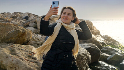 Young woman taking photos or herself at the winter sea using her smartphone while standing on the rocky shore. Perfect for travel and vacation footage.