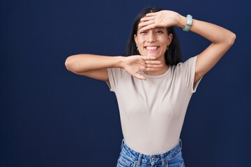 Young hispanic woman standing over blue background smiling cheerful playing peek a boo with hands...