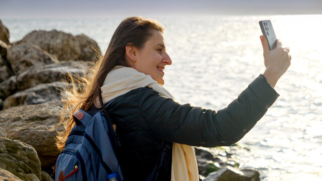 Smiling woman hiking and taking pictures of the stormy winter sea at sunset using her smartphone while standing on the rocks. Perfect for travel and tourism
