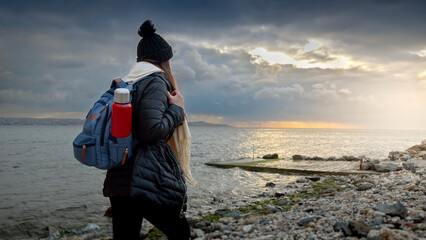 Young smiling woman with backpack and thermos walking along the rocky sea beach, enjoying the cool...