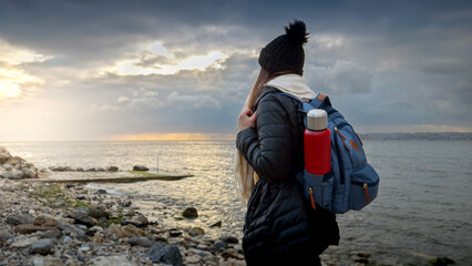 Female hiker in warm clothes with a backpack and thermos, walking on a rocky sea beach in cold, windy weather. Perfect for travel, tourism and adventure projects.