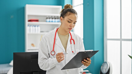 Young beautiful hispanic woman doctor reading document on clipboard with serious face at clinic
