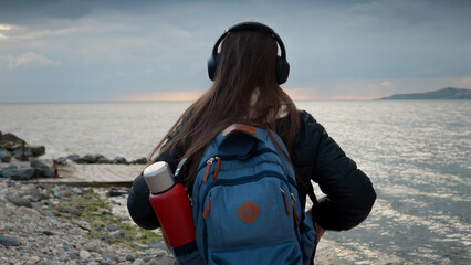 Smiling woman with backpack taking off headphones on sea beach and looks on calm cold ocean waves. Concept of travel, journey, enjoying music.