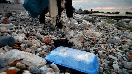 Fototapeta premium Young woman volunteer picking up trash on the beach with a plastic bag, showcasing her efforts to keep the environment clean and healthy.